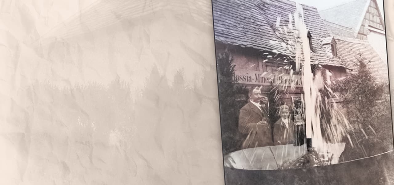 Historische Fotografie zweier Personen – ein Mann und ein Junge in formeller Kleidung – die vor einem Gebäude mit Fachwerkdach stehen. Im Vordergrund sprudelt ein hoher Wasserstrahl aus einem Brunnen. Über dem Eingang des Gebäudes ist der Schriftzug zu erkennen. Das Foto ist leicht verblasst und in eine beige strukturierte Hintergrundfläche eingebettet.