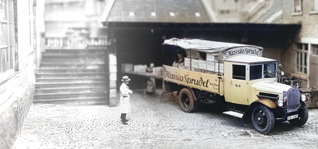 Historische Aufnahme eines Lieferwagens mit der Aufschrift ‚Hassia Sprudel‘, der in einem gepflasterten Hof vor einer überdachten Laderampe steht. Das Fahrzeug ist mit Kisten beladen und trägt die Nummer 4. Ein Mann im Mantel und Hut steht links im Bild und blickt zum Lastwagen. Die umliegenden Gebäude wirken alt und aus Backstein gebaut.