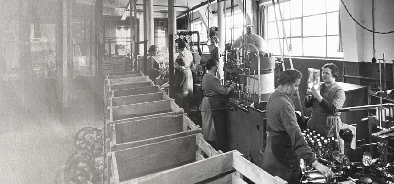 Schwarz-weiß-Fotografie einer Abfüllhalle, in der mehrere Arbeiterinnen und Arbeiter an einer Produktionslinie stehen. Sie befüllen Glasflaschen, kontrollieren sie im Licht und bedienen große, metallische Maschinen. Im Vordergrund stehen leere Holzkisten, im Hintergrund große Fenster, durch die Tageslicht in den Raum fällt.