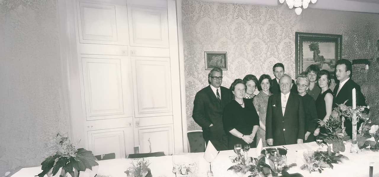 Schwarz-weißes Foto einer größeren Gruppe von Männern und Frauen, die eng zusammen vor einer reich gemusterten Tapete posieren. Sie stehen hinter einer festlich gedeckten Tafel mit Kerzen, Blumen und Gläsern. Die Personen wirken elegant gekleidet, als befänden sie sich auf einer Feier oder einem besonderen Anlass.