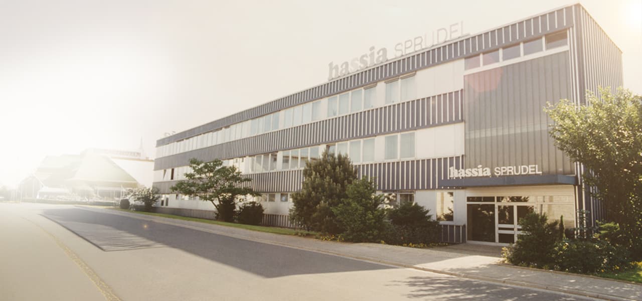 Farbaufnahme eines modernen Büro- und Produktionsgebäudes mit der Aufschrift ‚Hassia Sprudel‘ am Eingang und auf dem Dach. Das mehrstöckige Gebäude hat eine Fassade aus vertikalen Metallpaneelen und großen Fensterreihen. Davor stehen gepflegte Büsche und Bäume. Die Straße daneben ist leer und wirkt ruhig, das Bild ist von warmem Sonnenlicht überstrahlt.