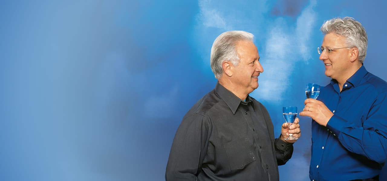 Zwei Männer stehen vor einem hellblauen Hintergrund und stoßen mit Wasser gefüllten Gläsern an. Beide lächeln sich an; der Mann links trägt ein dunkles Hemd, der Mann rechts ein blaues Hemd und eine Brille.