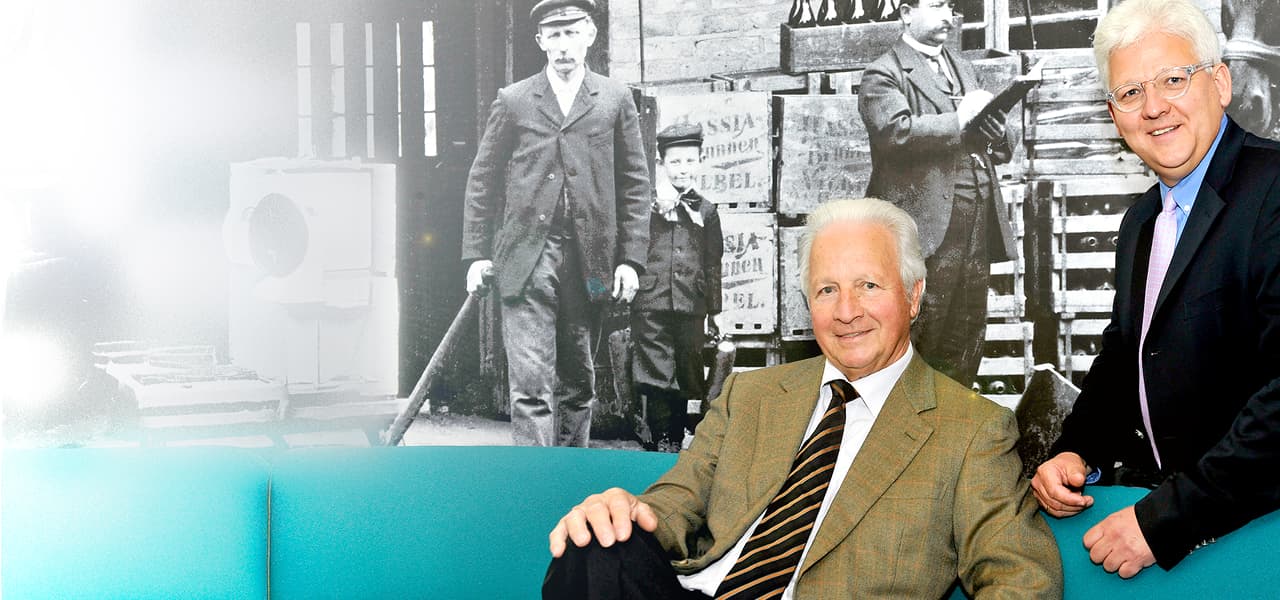 Zwei elegant gekleidete Männer sitzen bzw. stehen vor einer großen Wandgrafik in Schwarz-Weiß, die historische Arbeiter vor gestapelten Getränkekisten zeigt. Die beiden Männer im Vordergrund tragen Anzug und lächeln in die Kamera; einer sitzt auf einem türkisfarbenen Sofa, der andere lehnt daran. Die Bildkomposition verbindet moderne Farbaufnahme mit historischem Fotomotiv im Hintergrund.