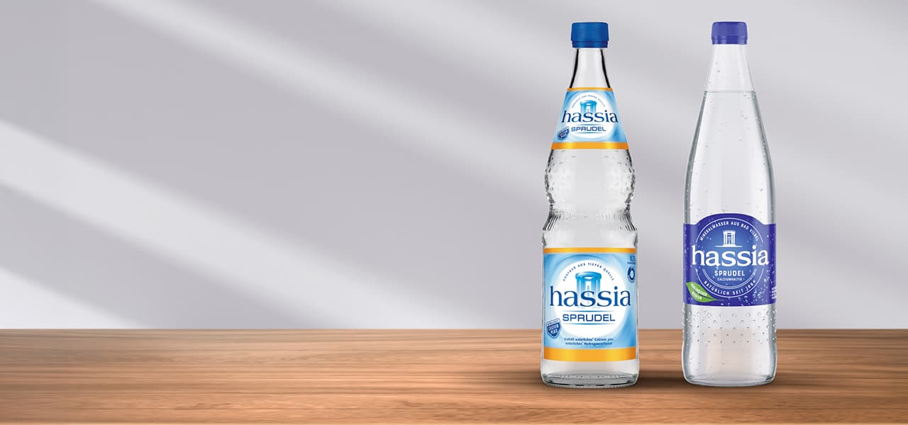 Zwei Glasflaschen Mineralwasser der Marke ‚hassia Sprudel‘ stehen auf einer Holzfläche vor einem hellgrauen Hintergrund. Links eine bauchigere Flasche mit blau-gelbem Etikett und blauem Deckel, rechts eine schlankere Flasche mit blau-violettem Etikett und lila Deckel. Beide wirken klar, frisch und modern inszeniert.