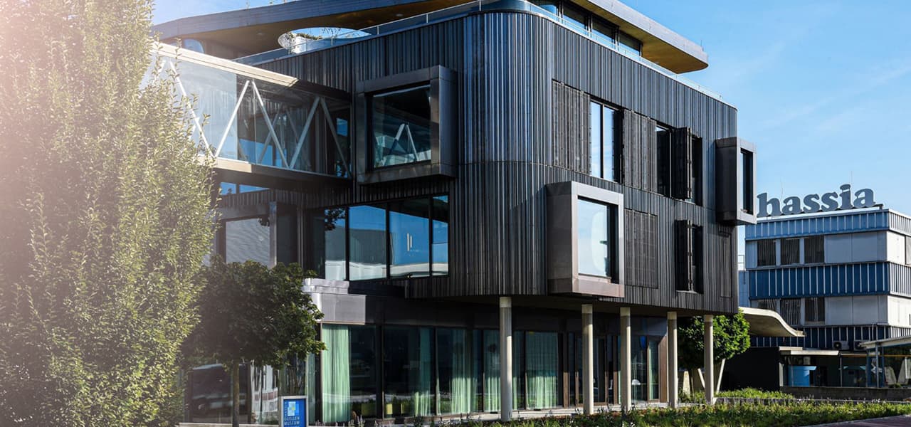 Moderne Firmengebäude mit markanter Architektur: Im Vordergrund ein mehrstöckiges Gebäude mit dunkler, vertikaler Fassadenstruktur, großen Fenstern und auskragenden Rahmen. Daneben Grünflächen und Bäume. Im Hintergrund ist ein weiteres Gebäude mit dem Schriftzug ‚hassia‘ auf dem Dach zu sehen. Die Szene wirkt sonnig und klar.